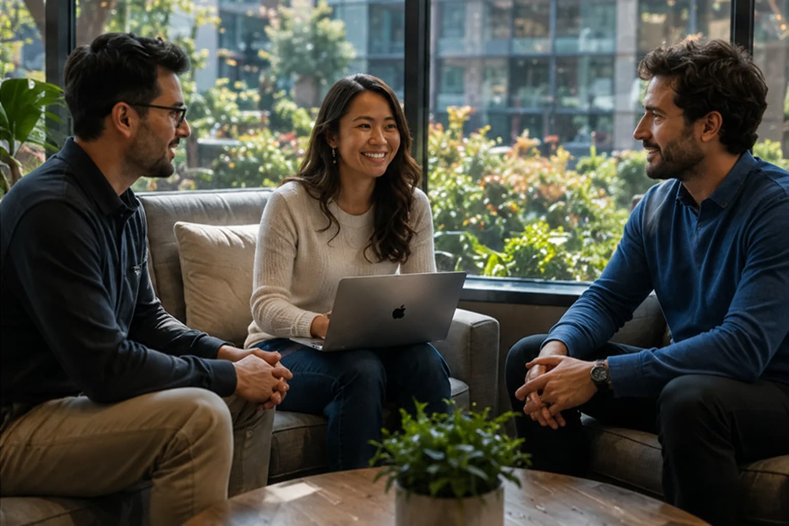 Three colleagues talking through a rollout plan on a laptop while seated on a sofa in a bright garden-facing lounge